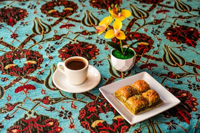 White cup of Turkish coffee on a saucer with a square plate of pistachio baklava and a small potted yellow orchid on a vibrant turquoise Ottoman-style patterned tablecloth.