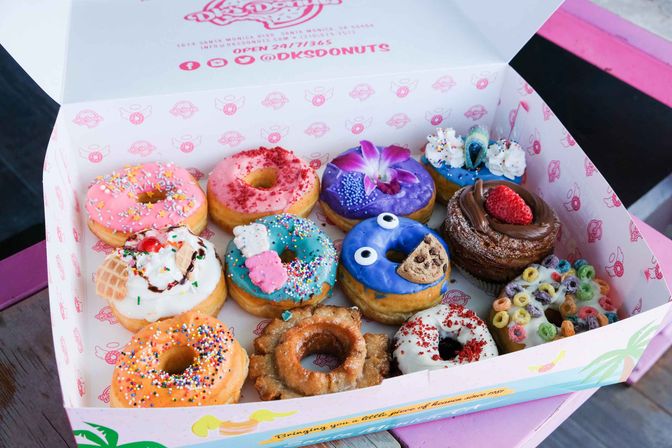 Assorted dozen colorful gourmet donuts in an open pink bakery box on a wooden table — glazed, sprinkled, cream-filled, chocolate- and fruit-topped varieties with cereal and novelty decorations