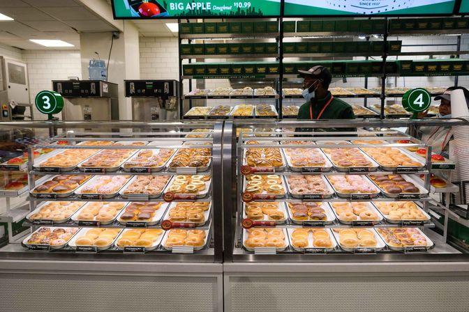 Glass bakery display case filled with trays of assorted donuts — glazed, chocolate, frosted and sprinkled — at a numbered in-store service counter with masked staff behind the counter.