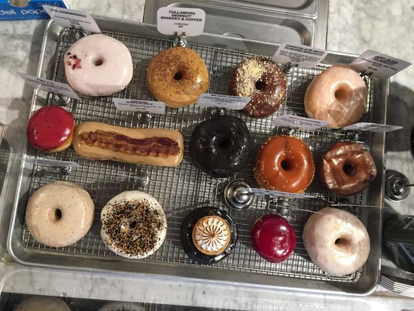 Colorful assortment of gourmet donuts — glazed, chocolate, caramel, maple-bacon and fruit-topped — arranged on a wire tray in a bakery display on a marble countertop.