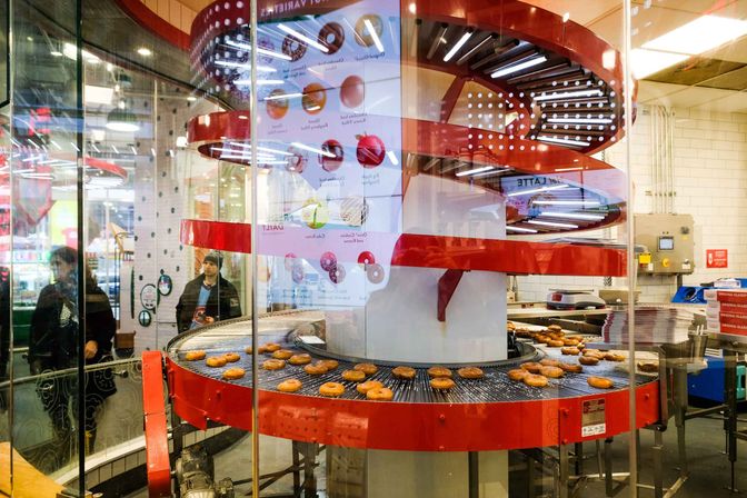Glass-front donut shop production line with a red spiral conveyor carrying fresh glazed donuts as customers watch through the storefront window