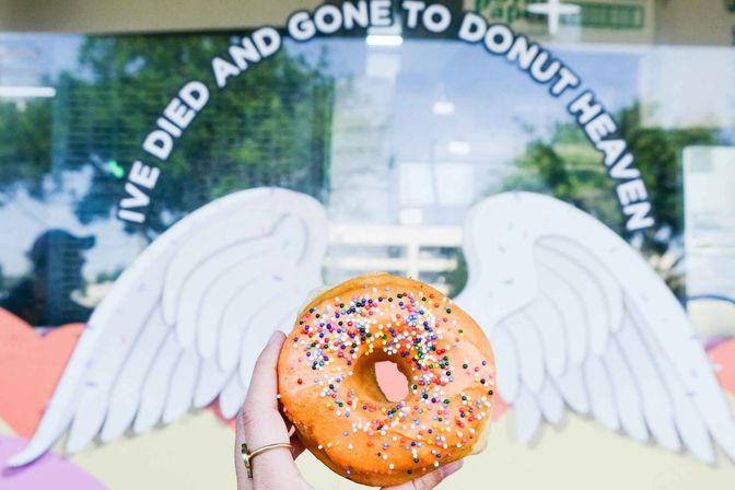 Hand holding a bright orange glazed donut with rainbow sprinkles against a storefront mural of angel wings and playful donut-themed lettering.