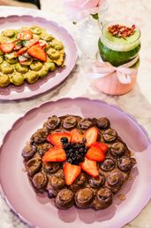 Chocolate bubble waffle topped with sliced strawberries and blueberries on a scalloped pink plate, paired with a green matcha-style drink garnished with dried rose petals and a pastel ribbon, with a green bubble waffle on a pink plate in the background.
