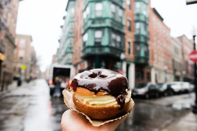 Hand holding a chocolate-glazed, cream-filled donut on a paper liner against a rainy city street with blurred brick buildings in the background.