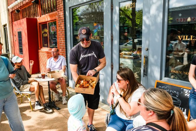 Smiling man in a baseball cap offers a box of assorted pastries to a child outside a busy downtown sidewalk bakery cafe with friends seated nearby.