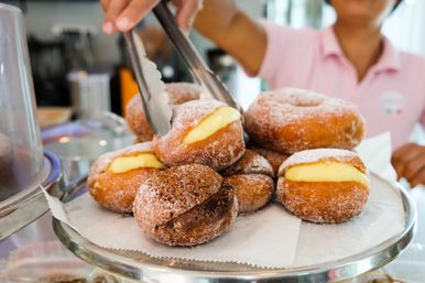 Pile of sugar-coated custard and chocolate-filled doughnuts on a glass cake stand, a hand with tongs picking one at a bakery counter