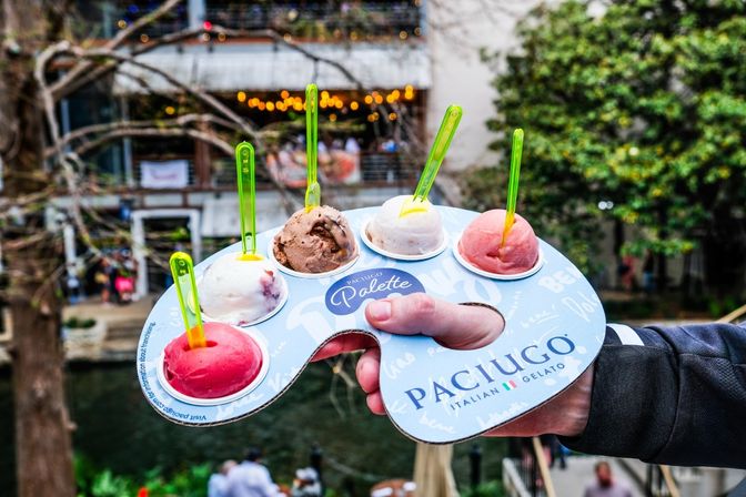 Hand holding a blue gelato tasting paddle with six colorful scoops and neon green spoons above a busy outdoor riverside walkway