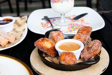 Cast-iron skillet of cinnamon-sugar churro bites with caramel dipping sauce on a wooden board, served on an outdoor café table with a sprinkle-topped milkshake and chocolate-dipped churros in the background.