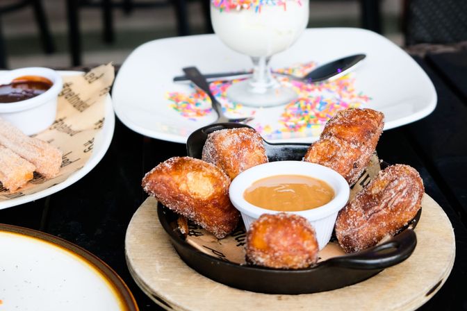 Cast-iron skillet of cinnamon-sugar churro bites with caramel dipping sauce on a wooden board, served on an outdoor café table with a sprinkle-topped milkshake and chocolate-dipped churros in the background.