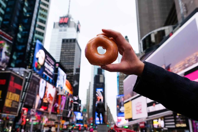 Glazed donut held up against bustling New York City Times Square with towering skyscrapers and colorful digital billboards