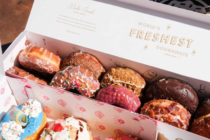 Sunlit bakery box of assorted artisan doughnuts — glazed, sprinkled, chocolate-covered, fruity and bacon-topped varieties in a pink-patterned box.