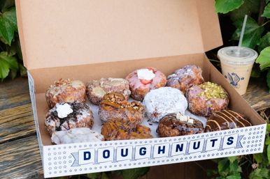 Open box of assorted gourmet doughnuts—powdered, glazed, chocolate- and pistachio-topped—next to iced coffee on a wooden bench with green foliage