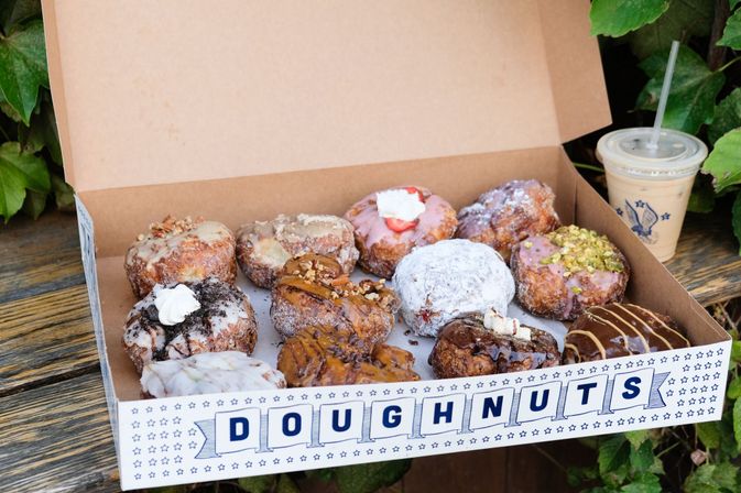 Open box of assorted gourmet doughnuts—powdered, glazed, chocolate- and pistachio-topped—next to iced coffee on a wooden bench with green foliage