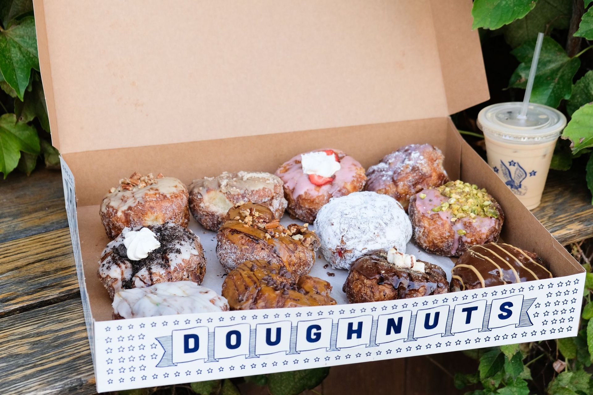 Open box of assorted gourmet doughnuts—powdered, glazed, chocolate- and pistachio-topped—next to iced coffee on a wooden bench with green foliage