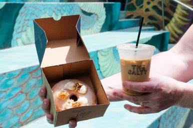 Hands hold a glazed donut with cream and crunchy topping in a brown takeout box and a clear plastic iced coffee with a black straw, set against colorful turquoise painted steps.