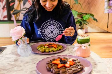 Person at a café table enjoying a green matcha bubble waffle topped with sliced strawberries, chocolate bubble waffle in foreground, pink rose in a vase and layered matcha drink with ribbon on a marble table.