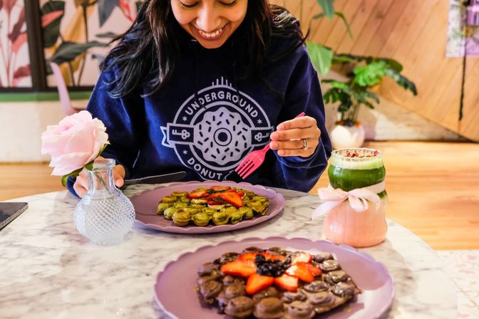 Person at a café table enjoying a green matcha bubble waffle topped with sliced strawberries, chocolate bubble waffle in foreground, pink rose in a vase and layered matcha drink with ribbon on a marble table.