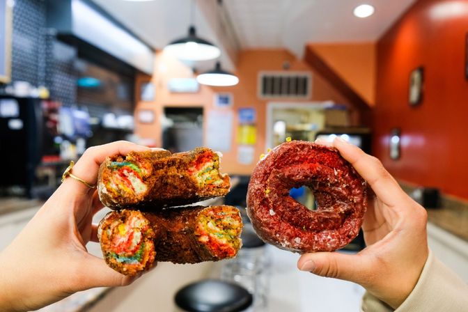 Two hands holding a sliced rainbow-filled cronut and a red glazed donut inside a bright orange-walled diner or bakery interior with counter seating and pendant lights