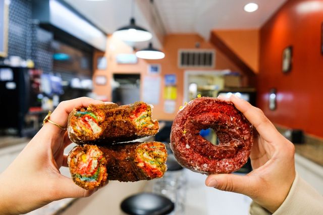 Two hands holding a sliced rainbow-filled cronut and a red glazed donut inside a bright orange-walled diner or bakery interior with counter seating and pendant lights