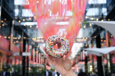 Hand holding a glazed donut with rainbow sprinkles in front of a vibrant downtown covered market installation with hanging pink ribbons and bokeh string lights.