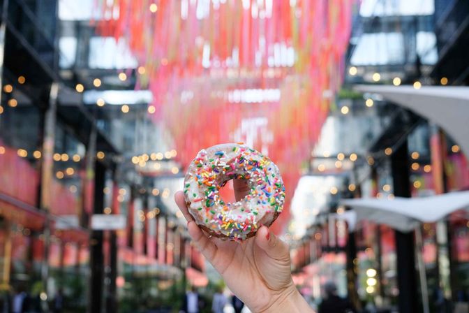 Hand holding a glazed donut with rainbow sprinkles in front of a vibrant downtown covered market installation with hanging pink ribbons and bokeh string lights.
