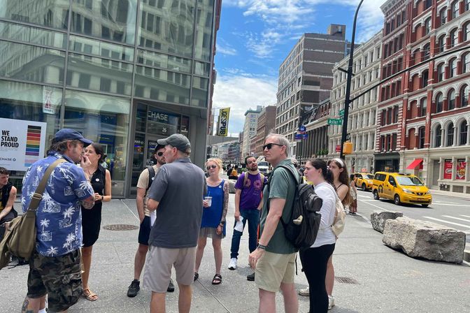 Group of people chatting on a sunny Astor Place sidewalk in Manhattan, with a glass-front building, cast-iron facades and yellow taxis passing by.