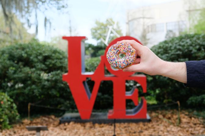 Hand holding a rainbow-sprinkled donut in front of a large red 'LOVE' sculpture in a leafy outdoor park with fallen leaves