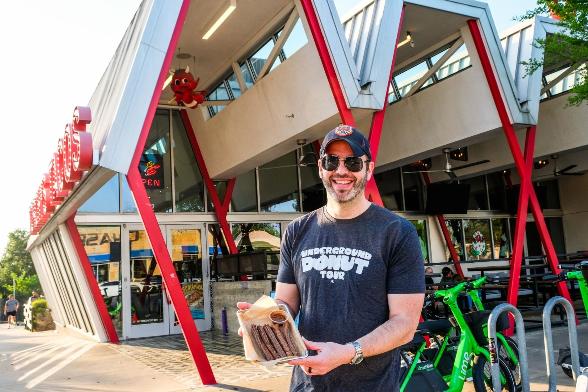 Smiling man in sunglasses and a cap holds a tray of churros with dipping sauce outside a retro red-and-white diner with angular supports and parked electric scooters on a sunny city sidewalk.