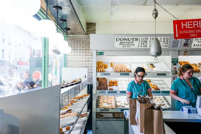 Bright city donut shop interior with glass-front display of assorted fresh donuts and pastries, two staff in teal uniforms packing paper bags at the counter while customers queue outside the window.