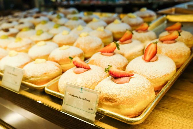 Bakery display of fresh sugar-dusted strawberry bombolones — round filled doughnuts topped with strawberry halves on gold trays.