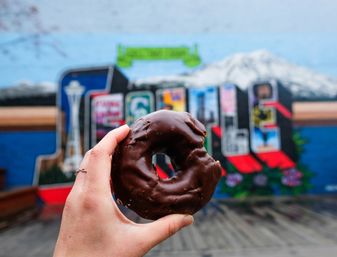 Hand holding a chocolate-glazed donut close to camera with a colorful Seattle mural showing the Space Needle and a snow-capped mountain blurred in the background
