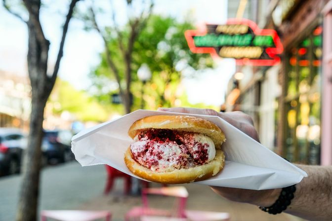 Close-up of a glazed doughnut ice cream sandwich held in a paper wrapper on a sunny downtown sidewalk, creamy vanilla ice cream with red crumbs and a blurred neon shop sign and trees in the background.