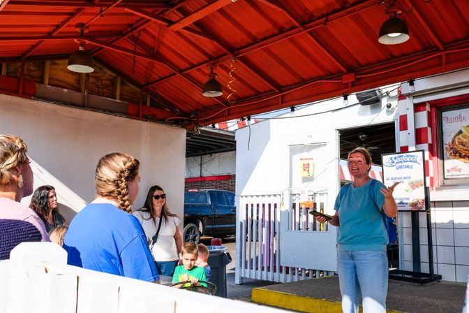 Sunny outdoor food stand with red metal awning and hanging lights, a staff member animatedly speaking to a small group of customers and children near a seafood specials sign.