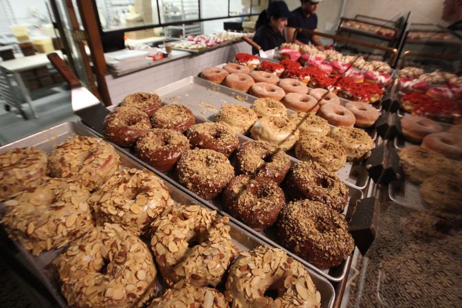 Assorted gourmet donuts on bakery trays — almond-crusted, chocolate crumble, glazed and powdered varieties in a bakery display case