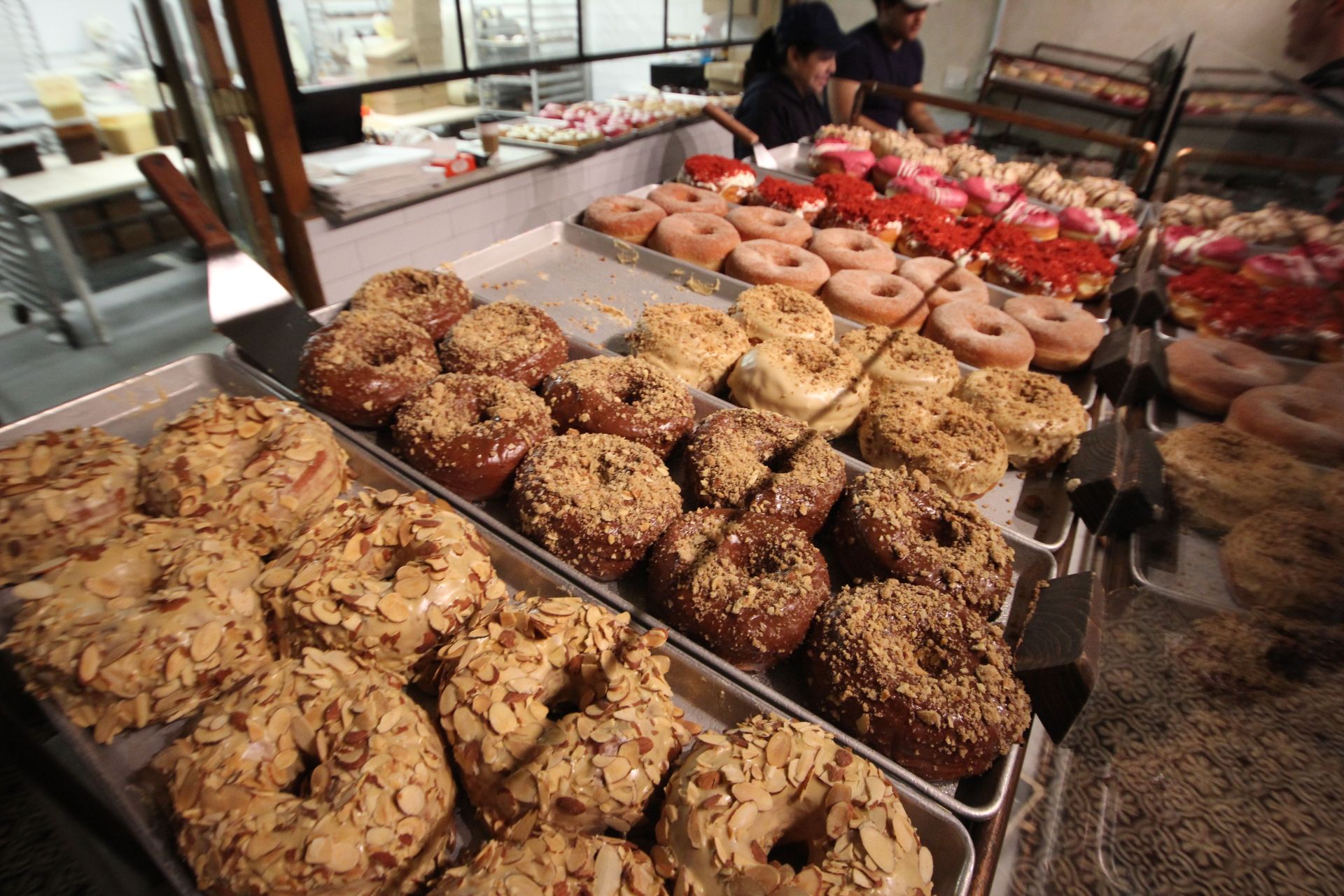 Assorted gourmet donuts on bakery trays — almond-crusted, chocolate crumble, glazed and powdered varieties in a bakery display case