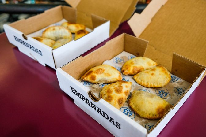 Six golden-brown baked empanadas with crimped edges in open takeaway boxes lined with paper on a maroon table