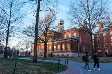 Red-brick college building with a white cupola on a crisp winter afternoon, leafless trees framing warm-lit arched windows and students walking along a curved campus path.