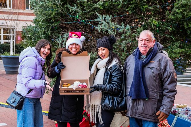 Smiling group of four in winter coats and a Santa hat pose by a large outdoor Christmas tree with string lights in a city plaza, holding an open box of assorted donuts.