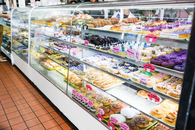 Glass bakery display case with rows of colorful assorted donuts and pastries topped with varied frostings and sprinkles