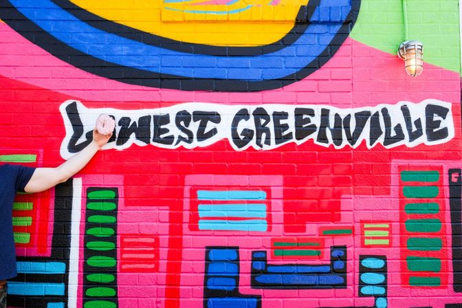 Vibrant Greenville street-art mural on a pink brick wall with bold graffiti lettering, colorful geometric shapes, and an outstretched arm holding a sprinkled pink donut.