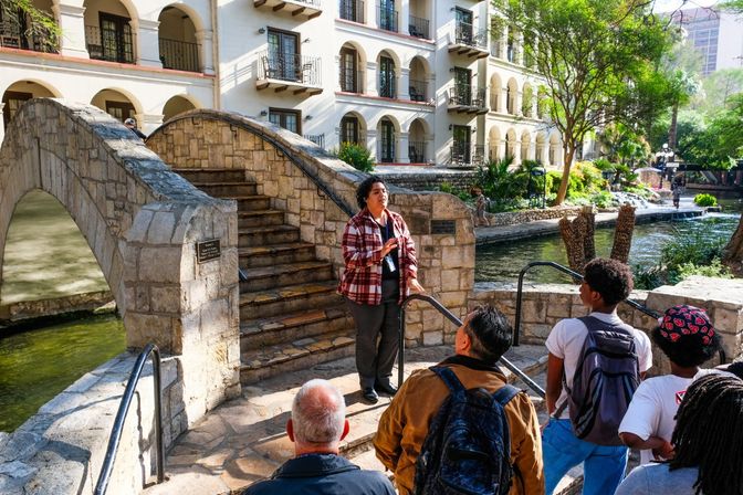 Tour guide addressing a small group by a curved stone footbridge and arched Spanish‑style building along the San Antonio River Walk, with river and trees in the background.