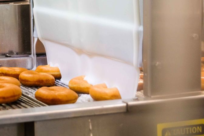 Fresh ring donuts on a conveyor belt being showered with white glaze from an industrial icing machine on a bakery production line