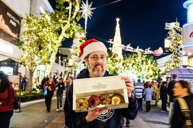 Person wearing a Santa hat smiles on a festive, tree-lined shopping street at night, holding an open box of assorted donuts with twinkling holiday lights, a decorated Christmas tree and seasonal street decorations in the background.