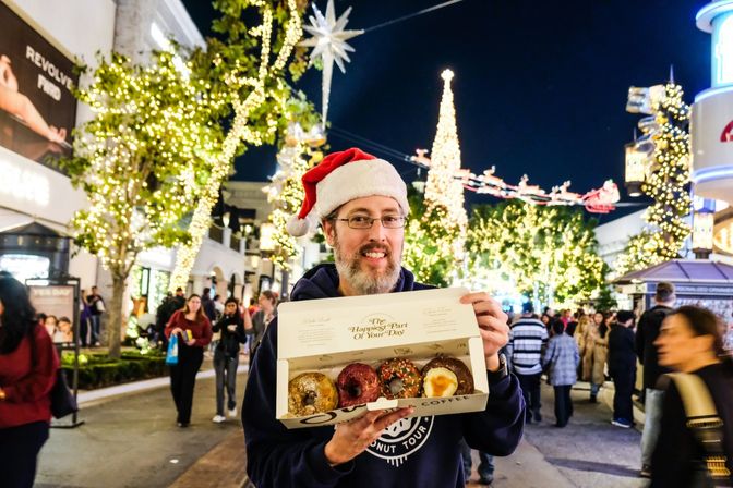 Person wearing a Santa hat smiles on a festive, tree-lined shopping street at night, holding an open box of assorted donuts with twinkling holiday lights, a decorated Christmas tree and seasonal street decorations in the background.