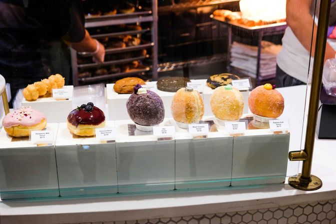 Glass bakery display case with colorful gourmet filled donuts — strawberry milk, coconut berry, ube, black sesame, pandan and passion fruit — bakers working behind the counter.