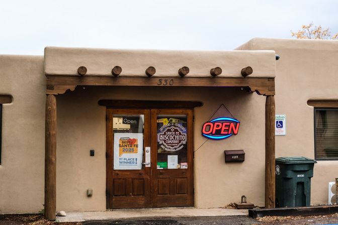 Adobe-style Santa Fe storefront with wooden double doors, exposed timber beams, a bright red-and-blue neon OPEN sign and an award poster on the door.