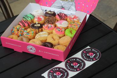 Pink box of assorted bite-sized mini donuts with colorful sprinkles, chocolate and cereal toppings on a black outdoor cafe table with round donut stickers beside it
