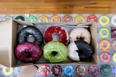 Overhead shot of six assorted colorful glazed gourmet donuts in a cardboard box held above a bakery counter with a patterned display of multicolored donuts