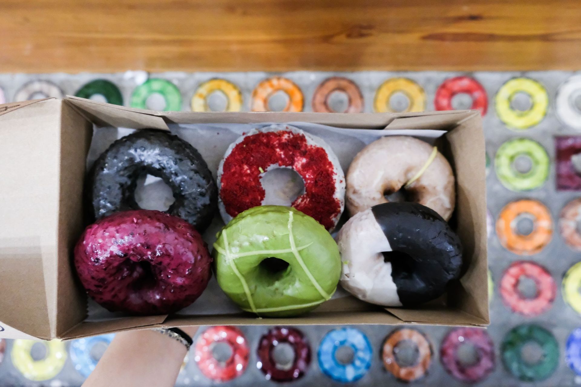 Overhead shot of six assorted colorful glazed gourmet donuts in a cardboard box held above a bakery counter with a patterned display of multicolored donuts