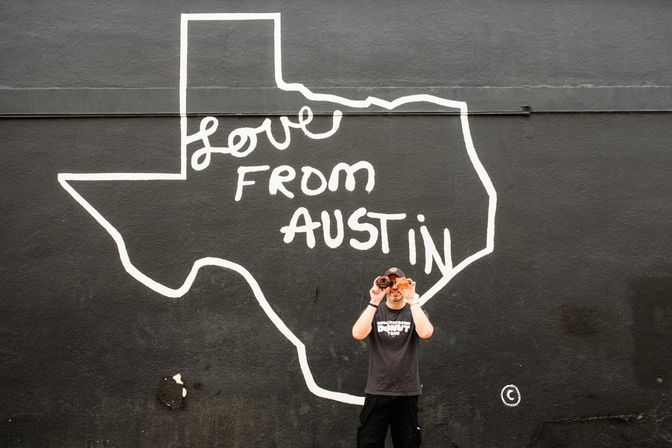 Person holding two donuts over their eyes in front of a black street mural of Texas outline reading Love From Austin — Austin, Texas street art